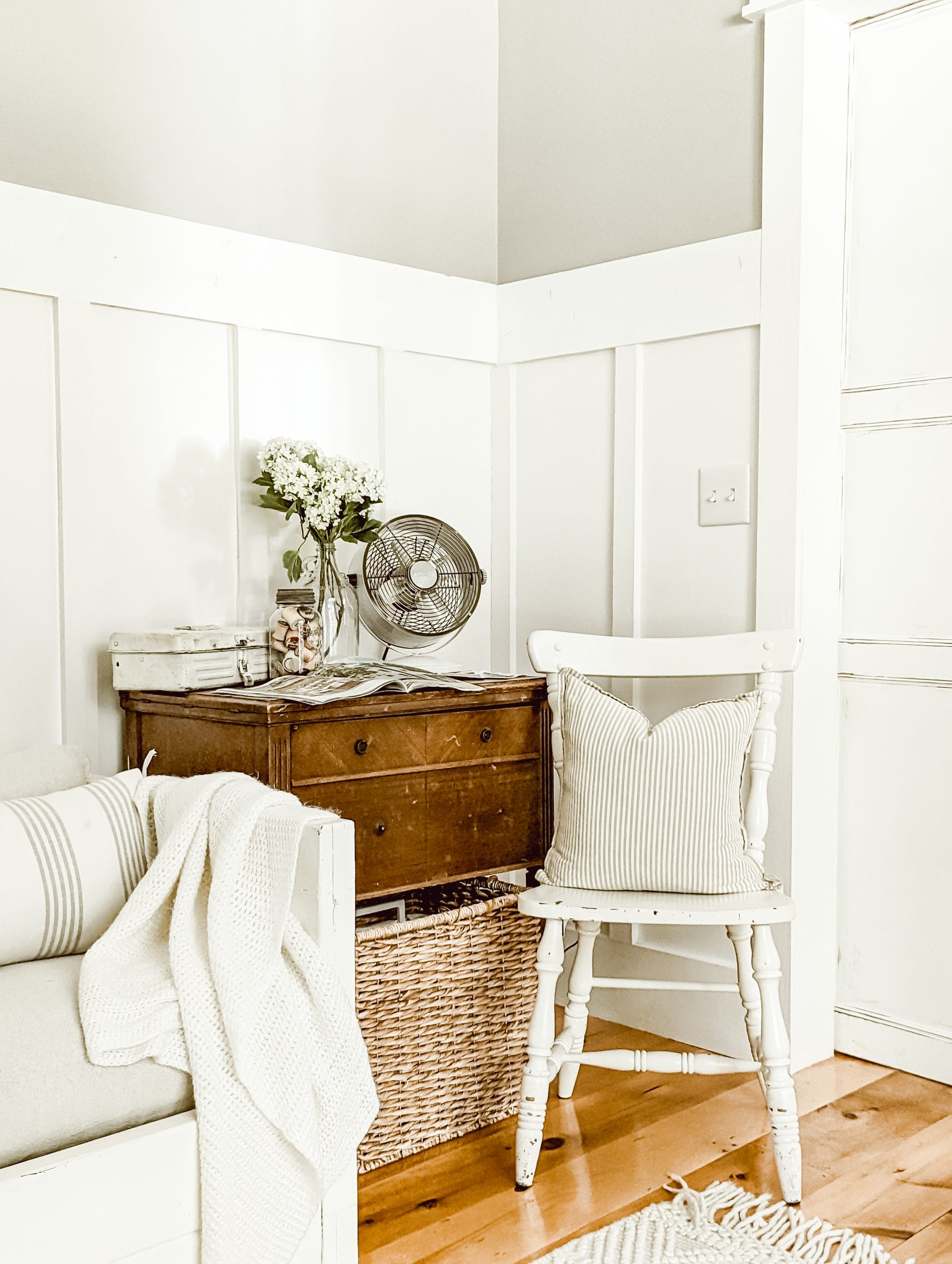 farmhouse cottage sitting area with white board and batten walls, chippy white furniture, and a wooden sewing cabinet as a side table